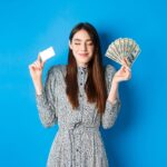 Medium shot of a model smiling with her eyes closed, holding up a fan of dollar bills and a business card. She is wearing a patterned dress and posing in front of a blue background.