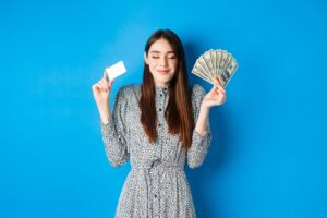Medium shot of a model smiling with her eyes closed, holding up a fan of dollar bills and a business card. She is wearing a patterned dress and posing in front of a blue background.